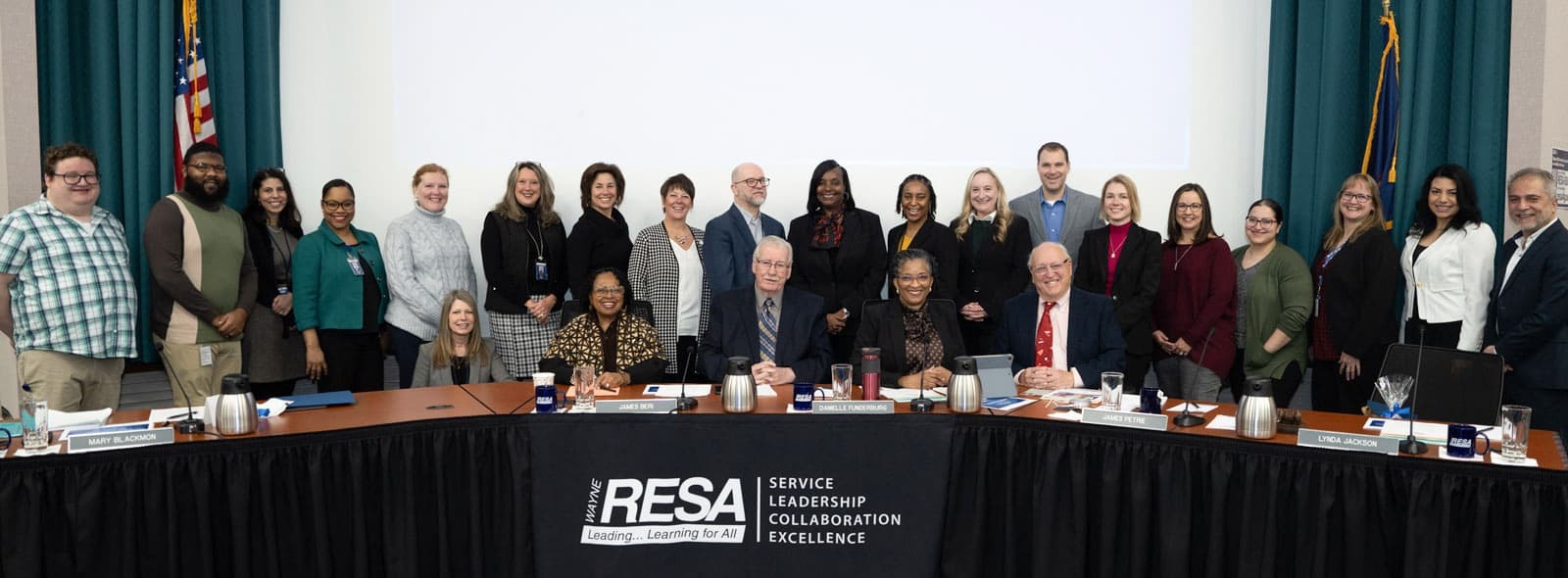 A diverse group of individuals gathered around a table, engaged in discussion, with a large screen displaying information behind them